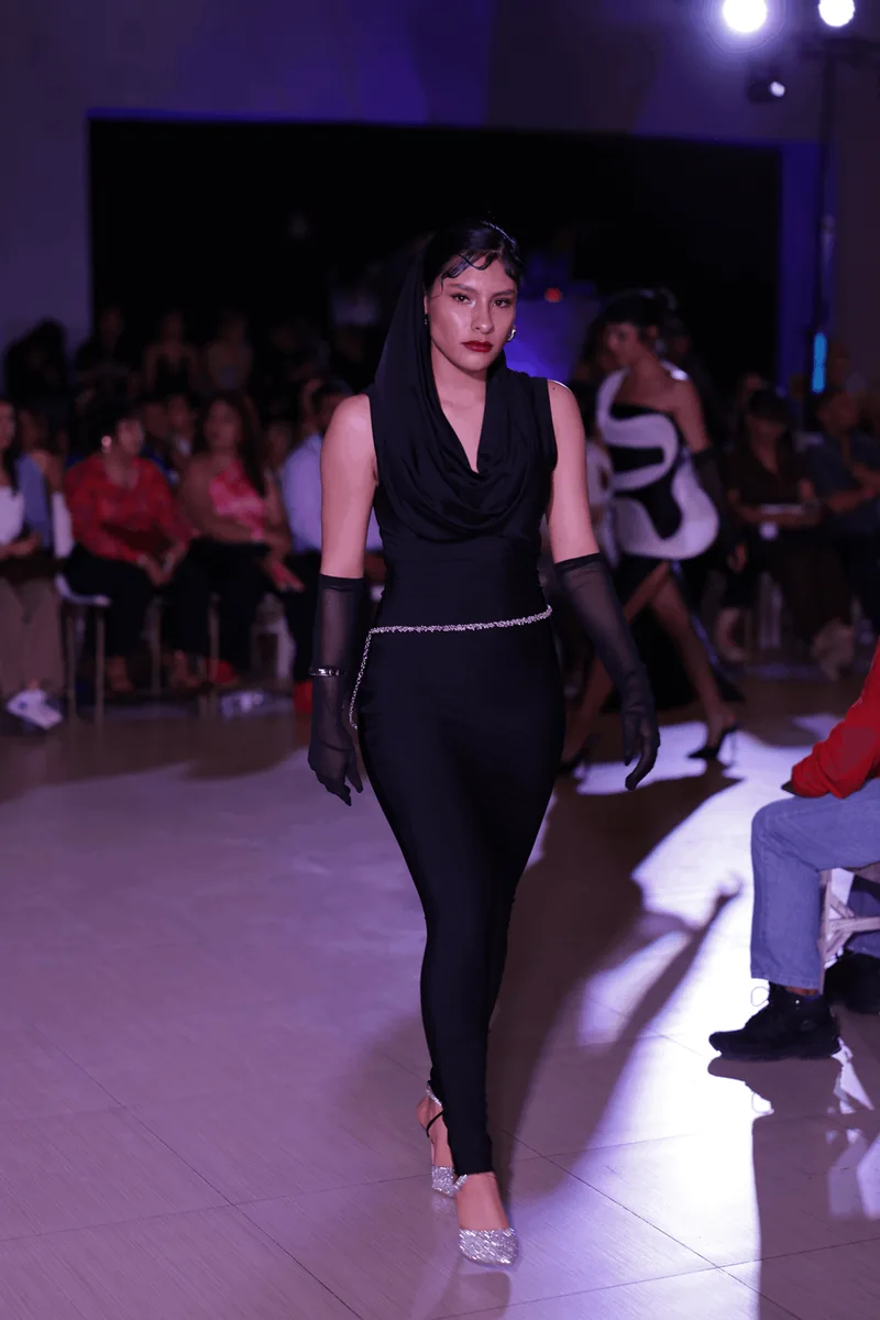 Vestido negro drapeado con capucha; segunda modelo al fondo.